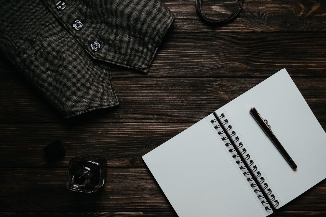 Top view of a rustic workspace with a notebook, pen, vest, and perfume bottle on a wooden table.